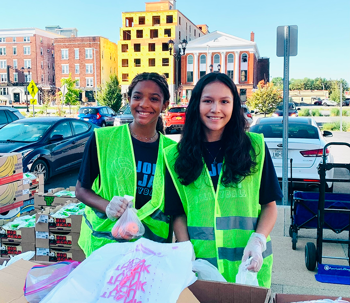 2 smiling people wearing green vests at a stand, parking lot and city buildings in the background